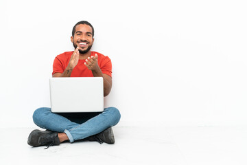 Young Ecuadorian man with a laptop sitting on the floor isolated on white background applauding...