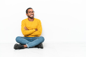 Young Ecuadorian man sitting on the floor isolated on white wall with arms crossed and looking forward