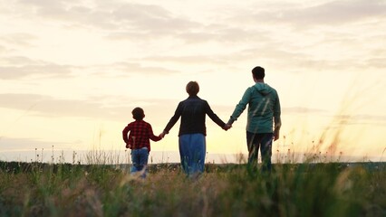 Happy family walks along grassy valley enjoying landscape at sunset. Sun rays fall on small family silhouettes walking along grassy valley. Family of parents and son walk along grass valley in evening