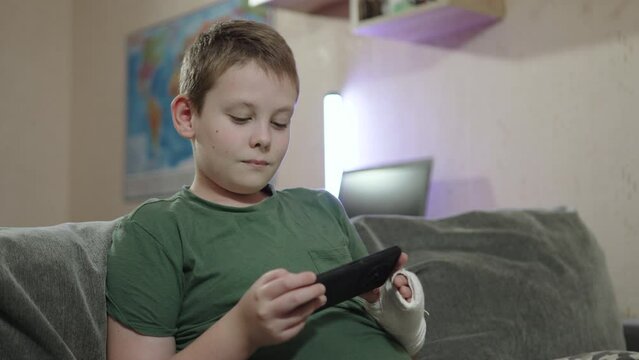 Attentive child with fracture and plaster on his arm watches video on smartphone on sofa. Watch educational online video about hand injury during recovery indoor. Distance learning during illness.
