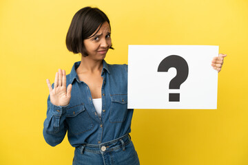 Young mixed race woman isolated on yellow background holding a placard with question mark symbol and doing stop sign