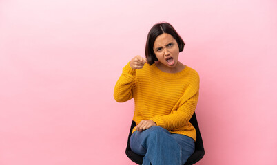 Young mixed race woman sitting on a chair isolated on pink background frustrated and pointing to...