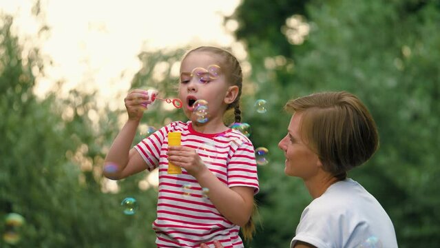 Mother and little girl find simple joy in leisurely activity of blowing soap bubbles among trees. Mom looks at daughter diligently blowing bubbles. Girl with braids blows bubbles supervised by mom