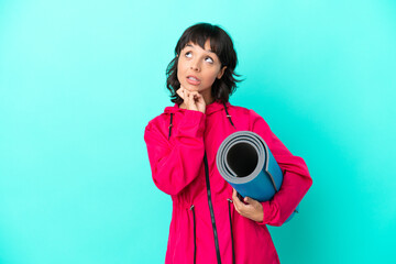 Young girl going to yoga classes while holding a mat isolated on blue background looking up while...