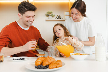 Obraz na płótnie Canvas Happy attractive family having breakfast sitting together in modern kitchen. Smiling father, mother, little cute daughter cooking, eating, drinking orange juice at cozy home. Healthy food concept 