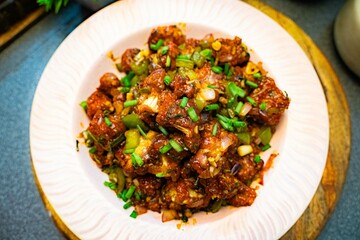 Veg Manchurian, Popular indo-chinese food made of cauliflower florets and other vegetable, served in a white plate over a rustic wooden table. selective focus. - obrazy, fototapety, plakaty