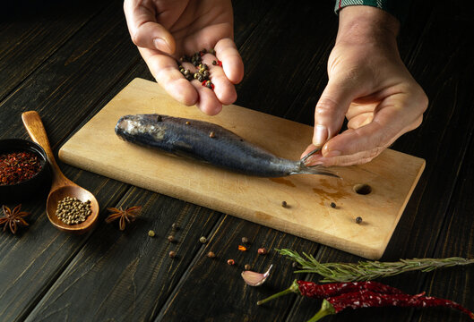 Close-up of hands adding dry pepper to Clupea fish. Concept of cooking pickled herring with spices on the kitchen table