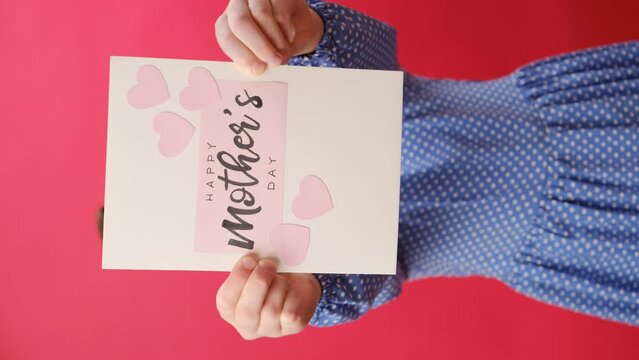 Vertical video of happy little girl kid holding card for Mother's Day with little pink hearts, posing isolated on plain red color background wall in studio. Spring holiday concept. Selective focus