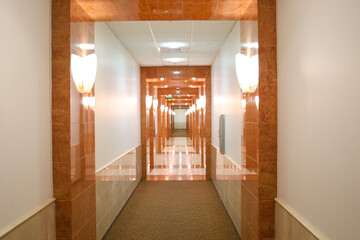 Long elegant hotel corridor white, beige and brown classical pattern on the walls. Clean, Sharp and straight hotel hallway. Interior design of walk way with wall lamp lights.