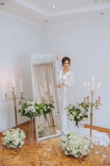 A beautiful brunette bride is preparing for the wedding in a beautiful boudoir style dress among fresh flowers and stylish candlesticks against a mirror background. Wedding portrait, photo.