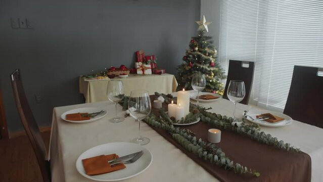 Handheld shot of Christmas table design with wine glasses, plates, cutlery and candles in cozy room with New Year tree and gifts in background
