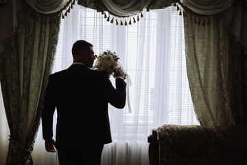 Stylish portrait of the groom with a bouquet of flowers in front of the window in the room. A man...