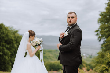 A handsome groom in the spring forest stands in front of his bride. Beautiful wedding white dress. Walks in the park. A happy and loving couple.