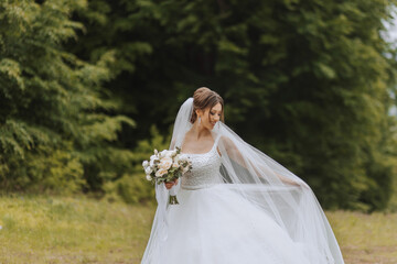 The bride in a lush dress and veil, holding a bouquet of white flowers and greenery, poses with the...