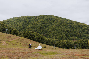 Portrait of a young bride and groom walking on green grass against the background of mountains after the wedding ceremony, side view. Happy wedding couple, copy space. Wide-angle photo.