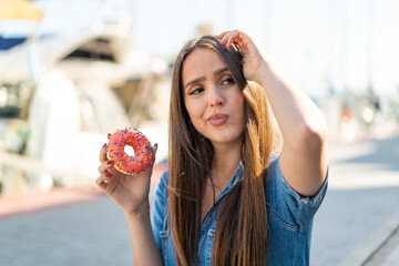 Young woman holding a donut at outdoors having doubts and with confuse face expression