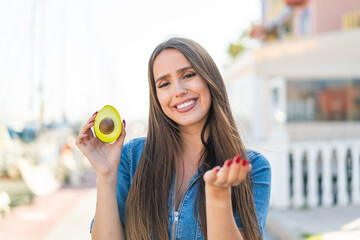 Young woman holding an avocado at outdoors inviting to come with hand. Happy that you came