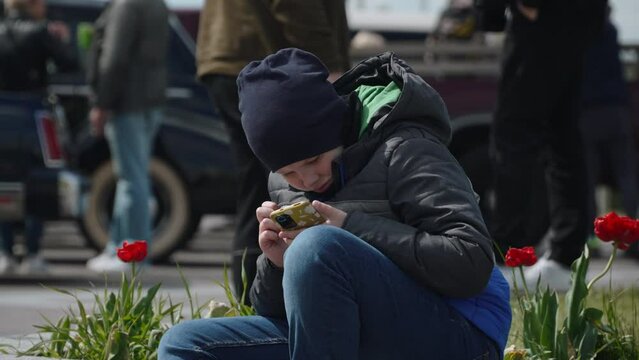 Boy with farsightedness looks closely at smartphone screen on street. Child with poor eyesight reads carefully while sitting on parapet of flower bed in city on cool spring day.