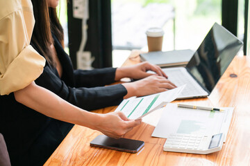 Two businesswomen read document and type on laptop while discussing marketing strategy of business