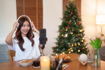 Young asian vj women wearing headphone and using digital smartphone to recording video and speaking morning talks about news in live streaming on channel while working with happiness at home studio
