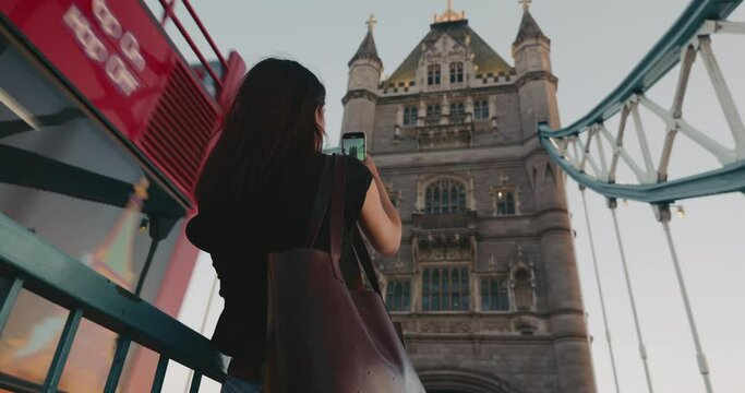 Young brunette female tourist taking picture of Tower bridge, using smartphone. View from the back.