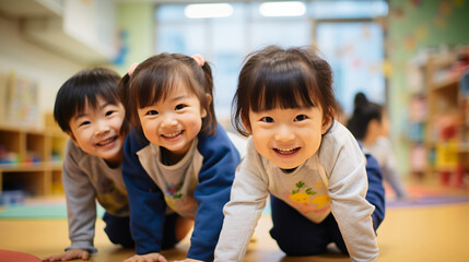 日本の幼稚園児・保育園児3人が長袖の私服で横に並んでハイハイでカメラに笑いながら向かってきている写真、背景教室、保育室のボケ - obrazy, fototapety, plakaty