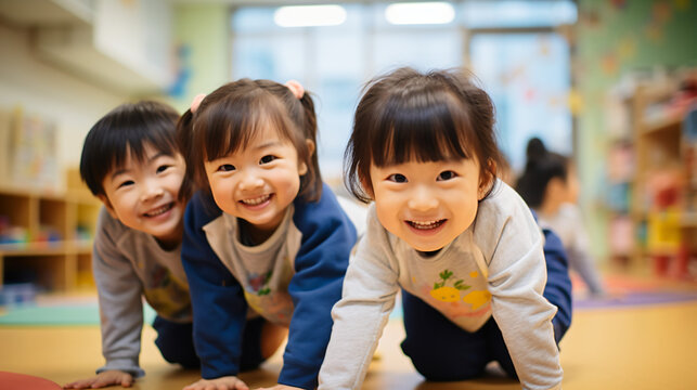 日本の幼稚園児・保育園児3人が長袖の私服で横に並んでハイハイでカメラに笑いながら向かってきている写真、背景教室、保育室のボケ