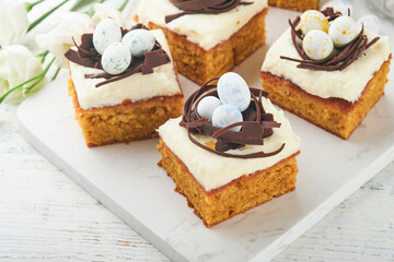 Easter carrot cake bars decorated with chocolate nest and chocolate candy eggs blossoming cherry or apple flowers on rustic light wooden backgrounds. Easter holiday meal. Traditional Easter food.