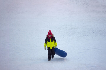 A little boy in warm bright outerwear carries a sled