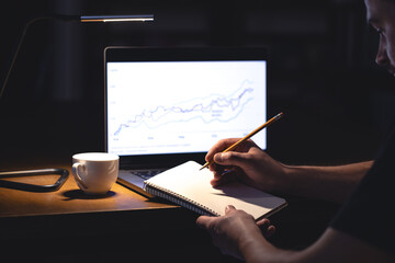 A man sits in front of a laptop with a chart and notepad at night.