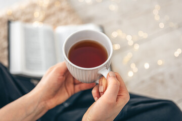 A cup of tea in female hands close-up, book on a carpet on the floor in a room.