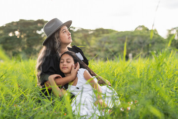 young latin mother with her daughter lying on her chest, sitting on the tall grass while the woman...
