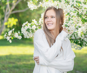 Face portrait of blonde young woman with cute smile wearing casual white blouse or shirt in the garden among blooming apple tree branches with small flowers and green leaves looking away at sunny day