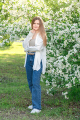 One blond young woman wearing white blouse and blue casual jeans looking straight at camera in the garden in front of blooming apple trees with small flowers and green leaves standing on grass lawn