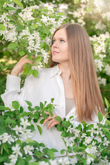Face portrait of blond young woman wearing casual white blouse or shirt in the garden among blooming apple tree branches with small flowers and green leaves standing with thoughtful smile looking away