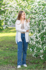 One cute blond young woman wearing white blouse or shirt and blue casual jeans in the garden in front of blooming apple trees with small flowers and green leaves standing on grass lawn thoughtful