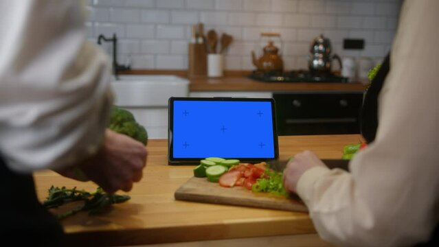 Tablet computer with mock up blue screen chroma key display on the kitchen counter, spouses chopping greens for salad or meal
