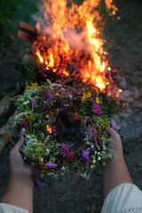 Girls hands hold floral wreath against bonfire, abstract dark natural background. Magic midsummer ritual. Summer Solstice Day. spiritual wiccan practice for Litha sabbat. witchcraft.