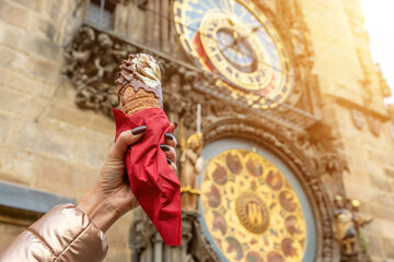 Female hand is holding traditional czech cookie trdelnik on background of Astronomical clock in Old Town of Prague city. - obrazy, fototapety, plakaty