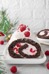Chocolate christmas roll cake with cream and fresh raspberries on a white plate on a gray background. Christmas yule log cake.