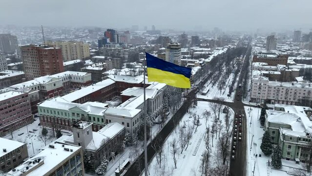 Fluttering on wind flag of Ukraine against background of winter Dnipro city and snowy street from a bird eye view during snowfall. Aerial drone view of roads and snow covered city. Ukrainian flag.