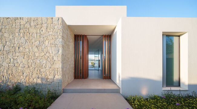 Contemporary house entryway featuring natural stone wall, wooden door, and clear sky in the morning light