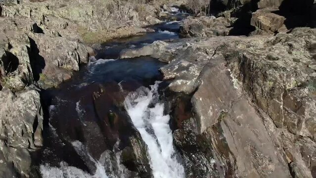 From above drone view of breathtaking scenery of rapid waterfall flowing through rocky boulder amidst leafless trees in Aljibe Waterfall, Guadalajara, Spain