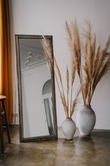 Elegant corner with a large mirror leaning against a wall and tall pampas grass in textured vases,...