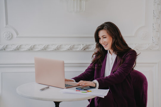 Professional woman in a purple blazer working happily on a laptop at an elegant white desk in a classic interior