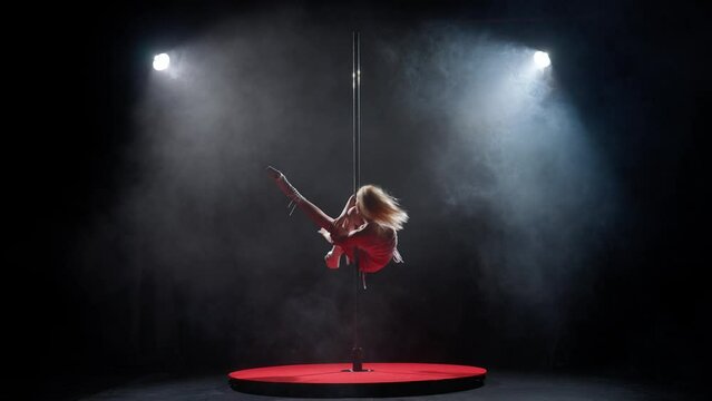Portrait of young female dancer in the studio. Professional pole dancer girl spinning on pylon, showing modern pole performance, black background with smoke.