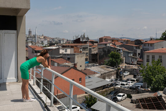 A young woman in a green dress leans on the railing of the terrace and looks into the distance at the poor panorama of Istanbul: Galata, Fatih districts.