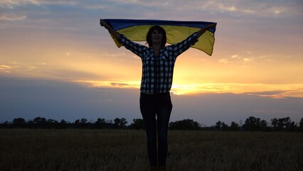 Happy lady walking on barley meadow with a raised over head flag of Ukraine. Ukrainian woman with a lifted blue-yellow banner on a beautiful sunset at background. End of war concept.