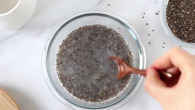 Hand mixing chia seeds and plant-based milk in a bowl to prepare vegan pudding. Top table view. Healthy breakfast food preparation concept.
