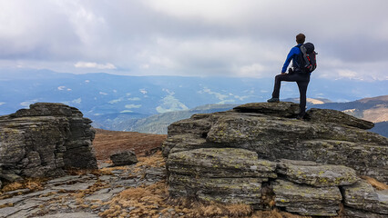 Man with backpack standing on massive rock formation at Steinerne Hochzeit, Saualpe, Lavanttal Alps, border Styria Carinthia, Austria, Europe. Panorama of alpine meadows and snowcapped mountains